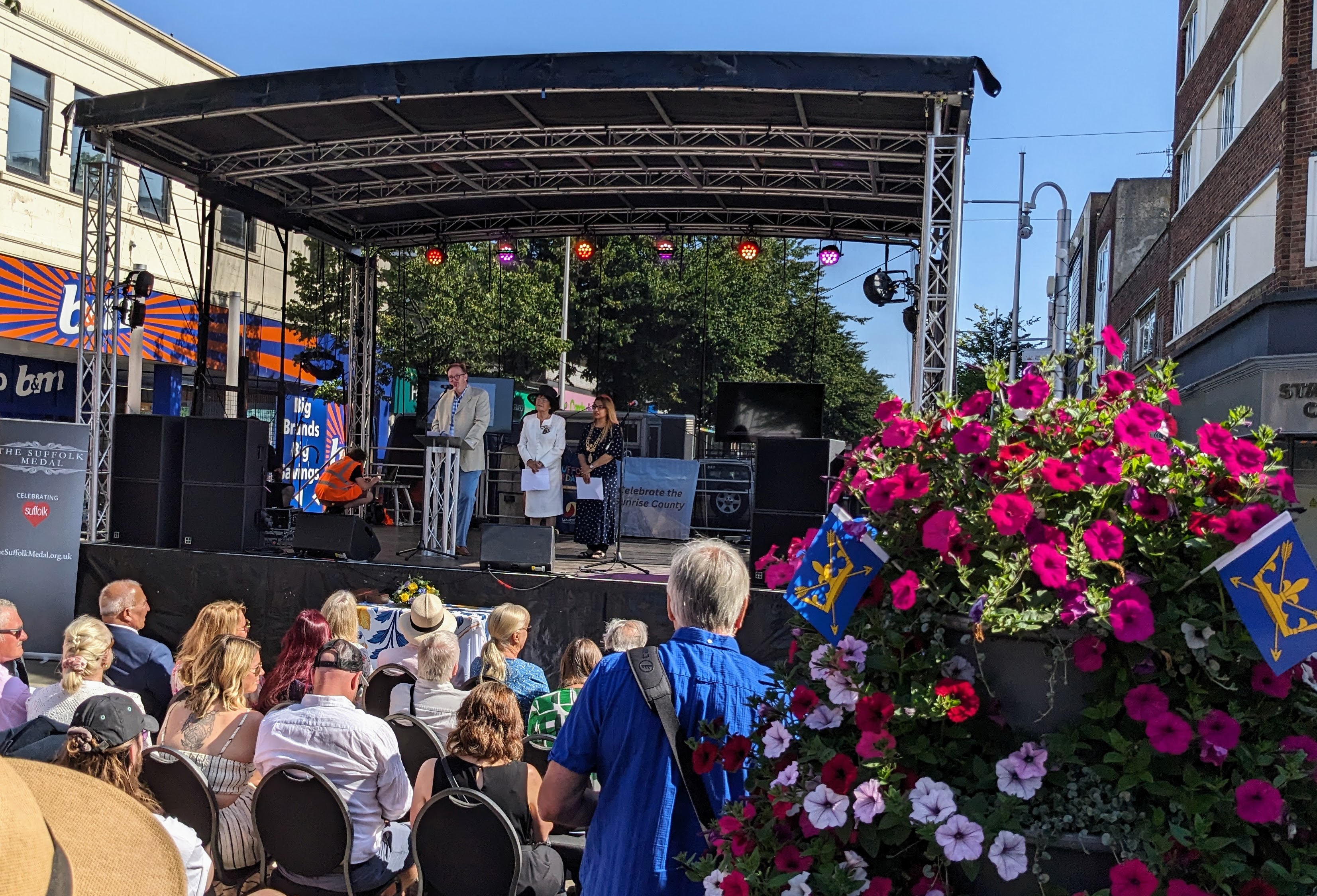 People on an outdoor stage with an audience, a planter with flowers in the foreground includes Suffolk Flags (blue background with a yellow crown and arrows through it)
