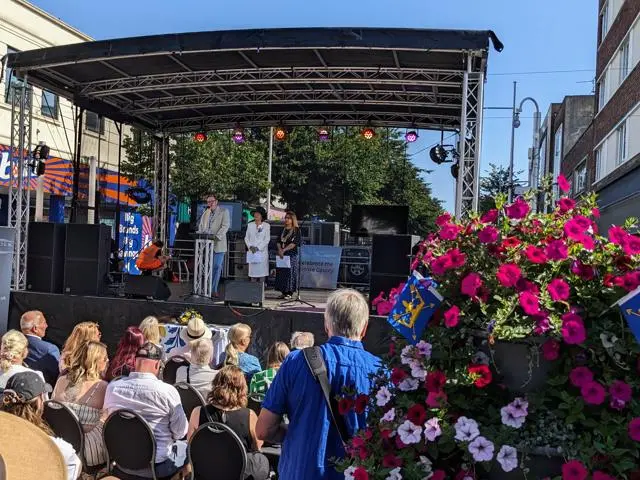 People on an outdoor stage with an audience, a planter with flowers in the foreground includes Suffolk Flags (blue background with a yellow crown and arrows through it)